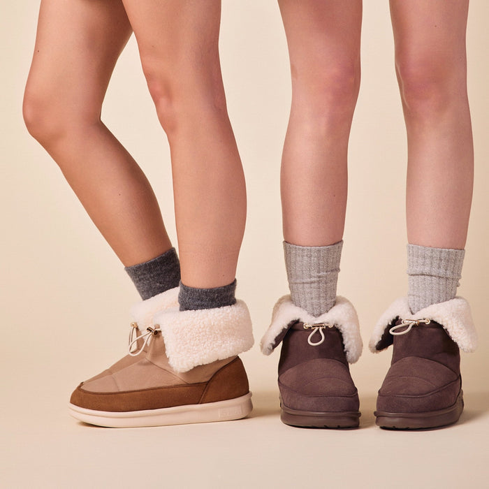 Three-quarter view of Apres Boot in camel nubuck on models' feet showcasing folded shearling cuff and low-profile sole on beige studio background