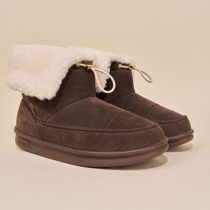 Three-quarter view of Apres Boot dark brown suede ankle boots with cream shearling cuff and toggle drawcord on beige background