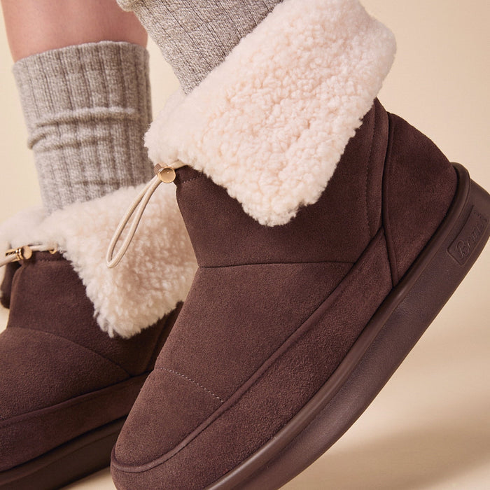 Side view of Apres Boot in dark brown suede with folded cream shearling cuff and molded rubber sole on beige background