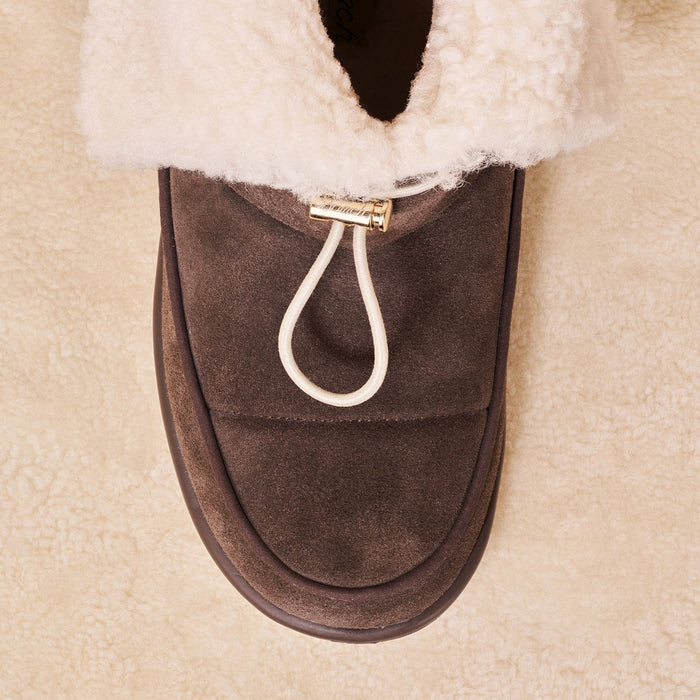 Top view of Apres Boot in dark brown suede showing folded shearling collar and front toggle loop on cream sherpa background
