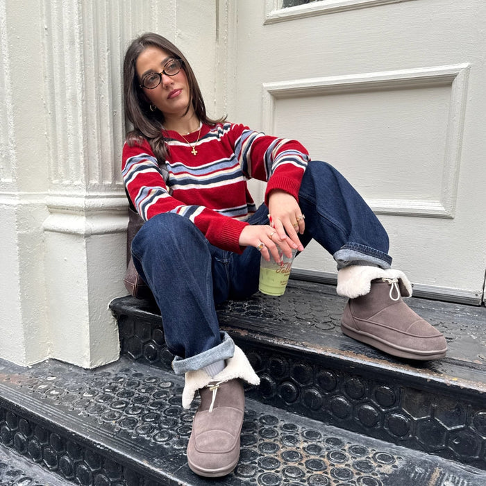 Woman sitting on outdoor steps wearing dark brown suede Apres boots with folded shearling cuff, holding an iced drink