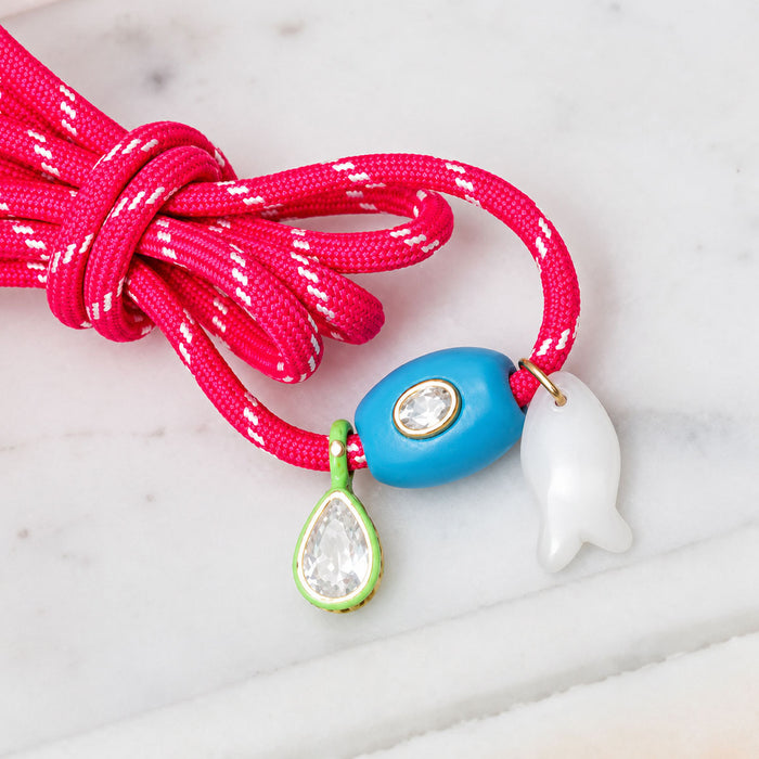 Close-up of Carrie Charm Kit coral-pink braided charm bracelet with blue oval bead, white fish charm, and green teardrop on light marble background
