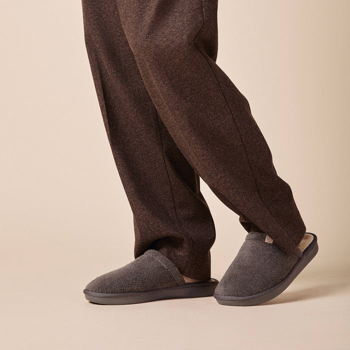 Side view of Essential slipper in dark grey terry showing closed-toe terry upper and cushioned EVA sole on beige studio floor