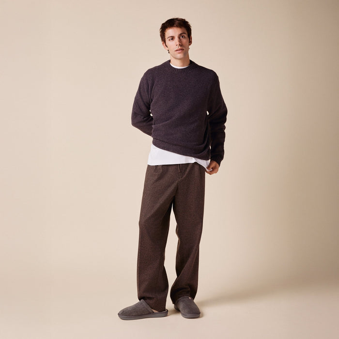 Man standing wearing dark grey terry slippers with relaxed trousers and a sweater on a beige studio background