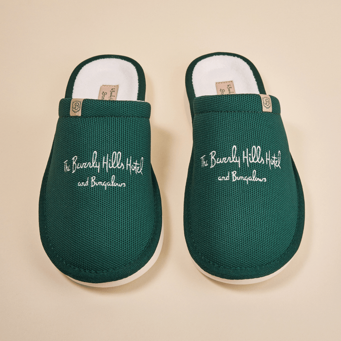 Top-down view of green Essential slippers embroidered with The Beverly Hills Hotel and Bungalows text on a beige background