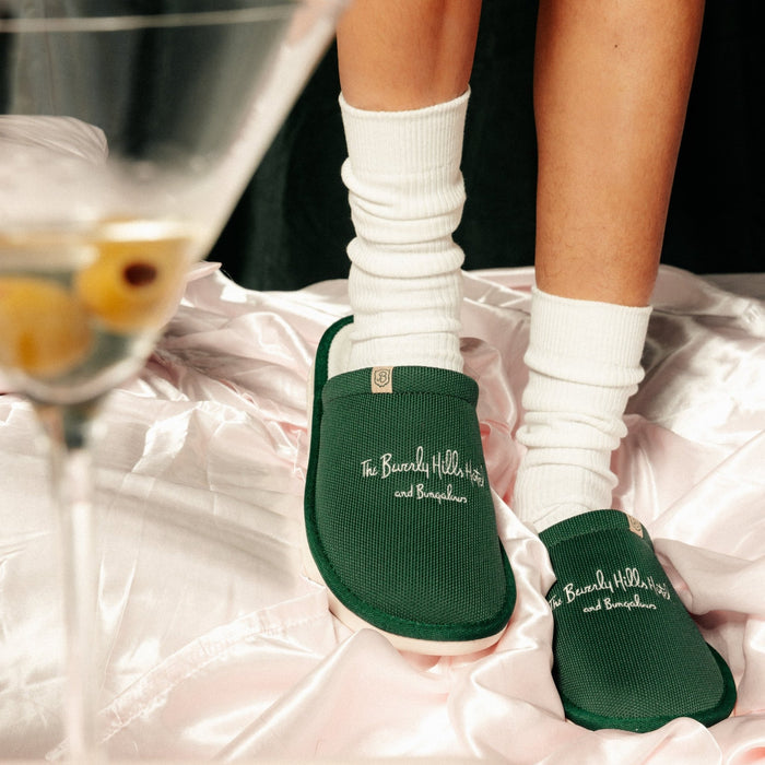 Front of green Essential Beverly Hills Hotel slippers with embroidered hotel logo on knit upper, worn with white socks on pink satin sheets