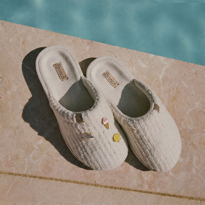 Angled top view of cream ribbed terry slippers showcasing three enamel charms on a stone poolside ledge