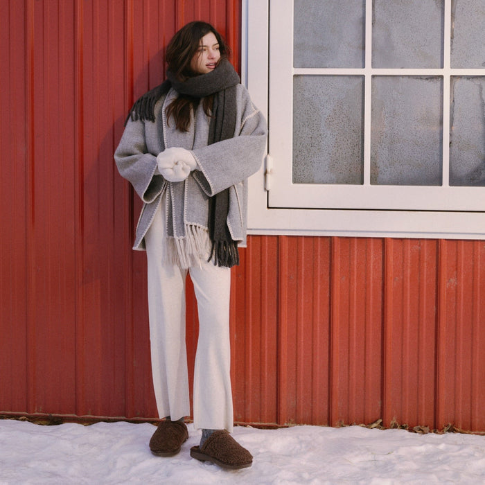 Woman standing outdoors wearing brown shearling slippers beside a red building with snow on the ground
