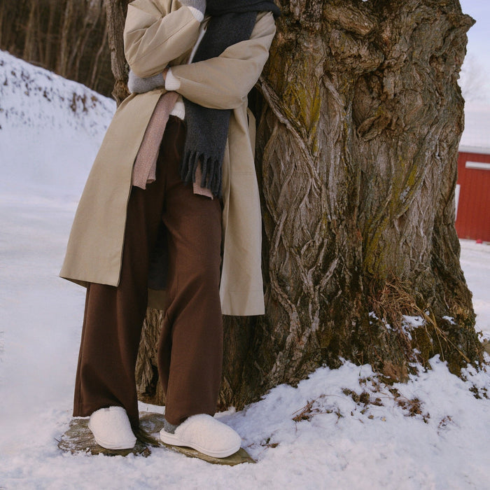 Model wearing natural all-shearling slip-on slippers standing on snow beside a large tree