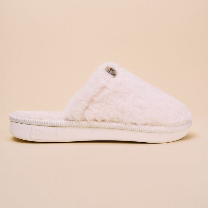 Side view of Essential All Shearling slipper showcasing plush natural shearling upper and cushioned white sole on beige background