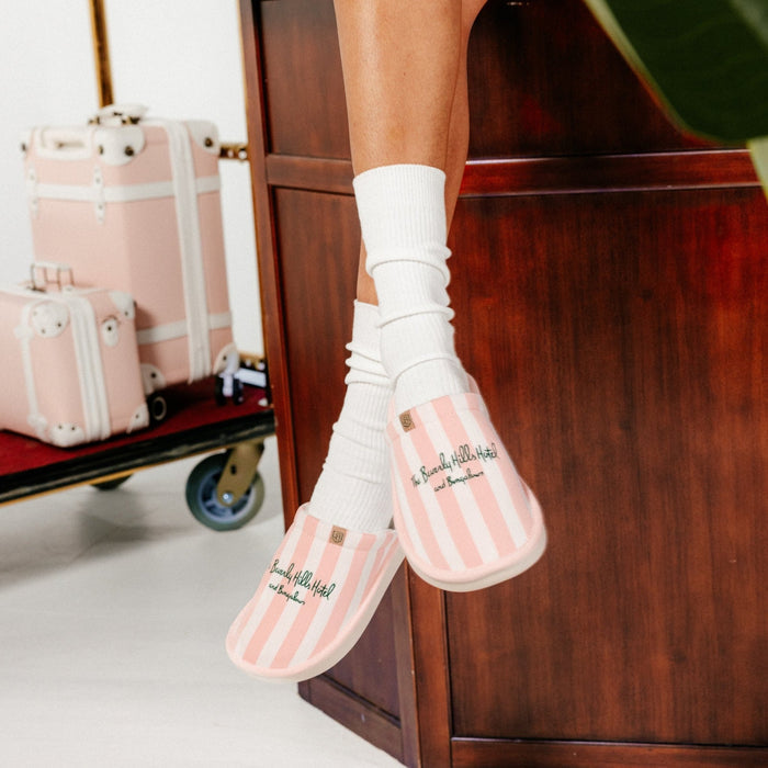 Angled view of pink-striped Essential slipper showing embroidered Beverly Hills Hotel logo and cushioned sole with model wearing white socks and wooden desk background