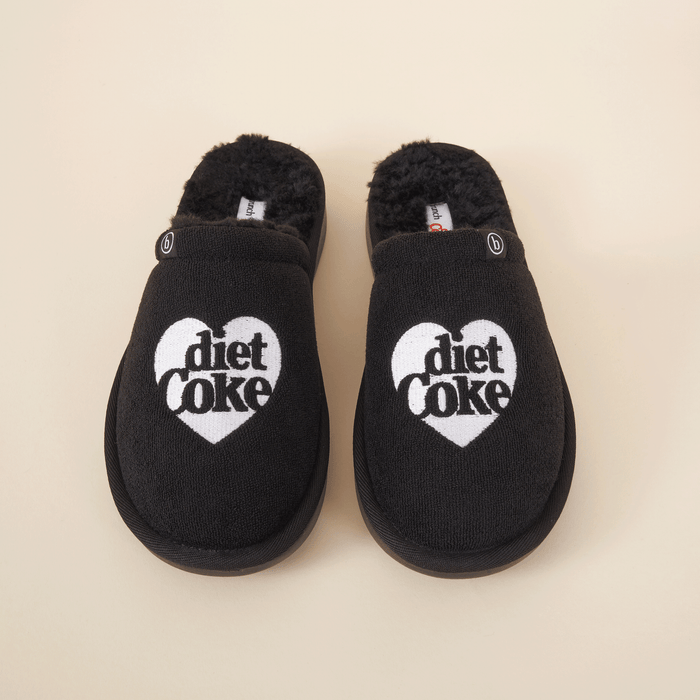 Top-down view of black shearling slippers showcasing white heart-shaped Diet Coke embroidery and faux-shearling lining on cream background