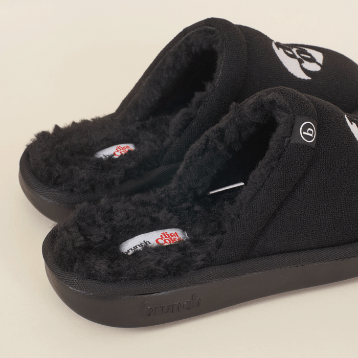 Three-quarter view of black slip-on slippers with faux shearling lining, Diet Coke footbed label and embossed Brunch sole