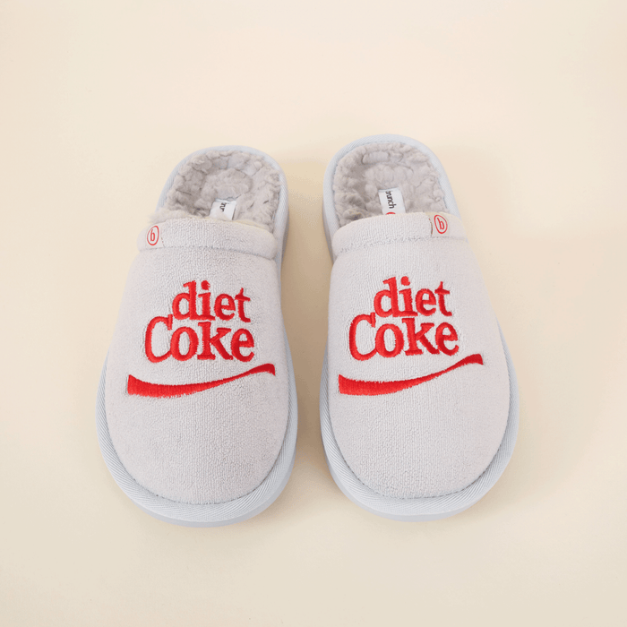 Top-down view of grey shearling slippers with red embroidered diet Coke logo on beige background
