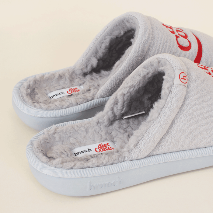 Angled close-up of grey Diet Coke x Brunch slippers with shearling lining and red embroidered logo on pale background