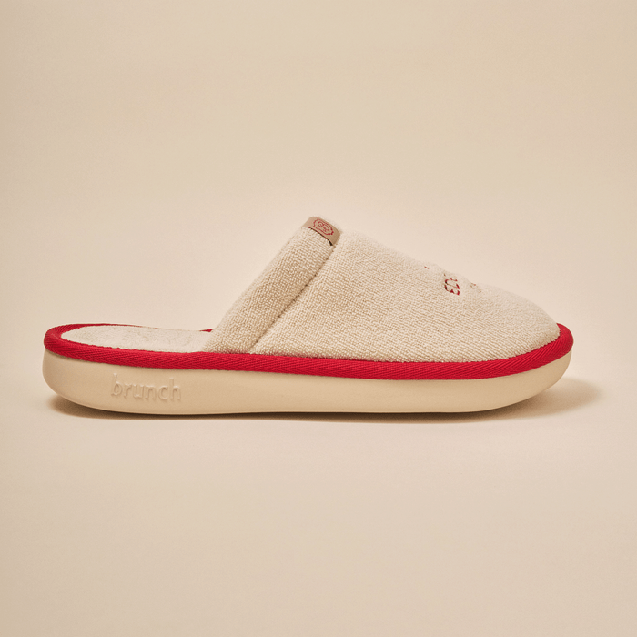 Side view of Essential Eden Rock slipper in beige with soft terry upper, red trim and Eden Roc embroidery on neutral backdrop