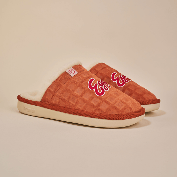 Three-quarter view of Essential Eggo slippers with waffle-embossed suede upper, Eggo embroidered logo, cream shearling lining on beige background