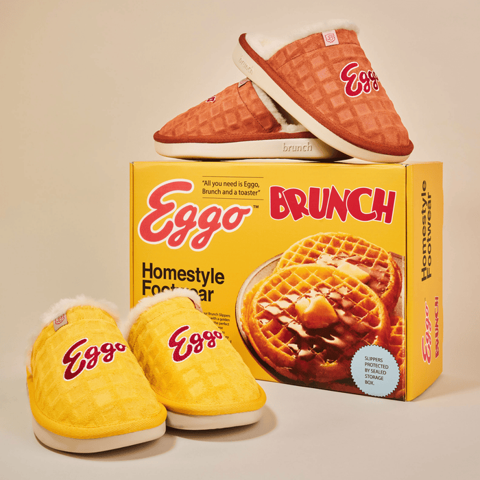 Front three-quarter view of yellow Essential Eggo slippers showing waffle‑embossed suede, shearling lining, embroidered Eggo logo, on Eggo Brunch box