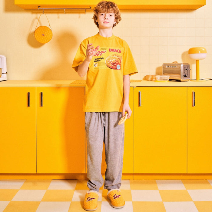 Person standing in a yellow kitchen wearing yellow Eggo slippers and a matching Eggo t-shirt while holding a waffle