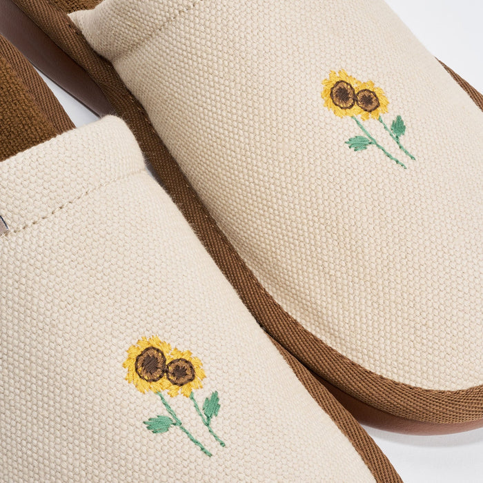 Close-up top view of beige canvas slipper showing hand-stitched yellow sunflower embroidery and brown trim on white background
