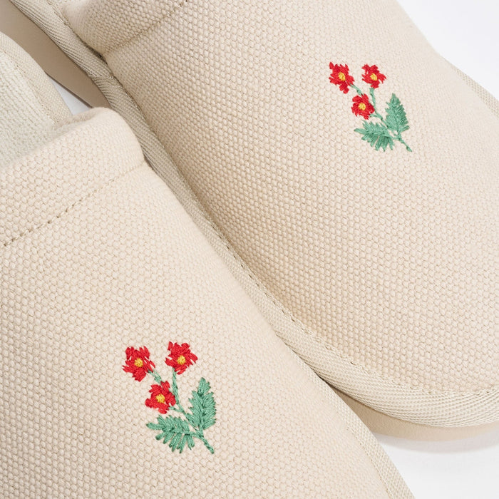 Top view of pair of beige canvas slippers showcasing hand stitched red wildflower embroidery on toes with white background