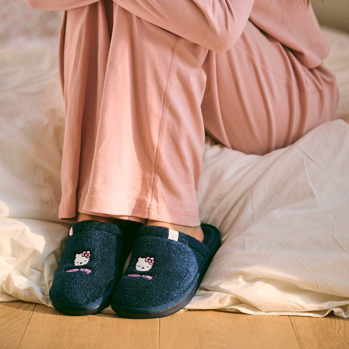 Front view of navy terry slip-on slippers with embroidered Hello Kitty, worn by a person sitting on a bed