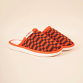 Three-quarter view of Amber knit checkered slip-on mule slipper showing orange-and-black knit upper, white cushioned sole, beige background