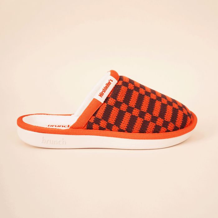 Side view of Brunch x Hirshleifers Essential slipper showing orange-and-black knit checker upper and white sole on beige background