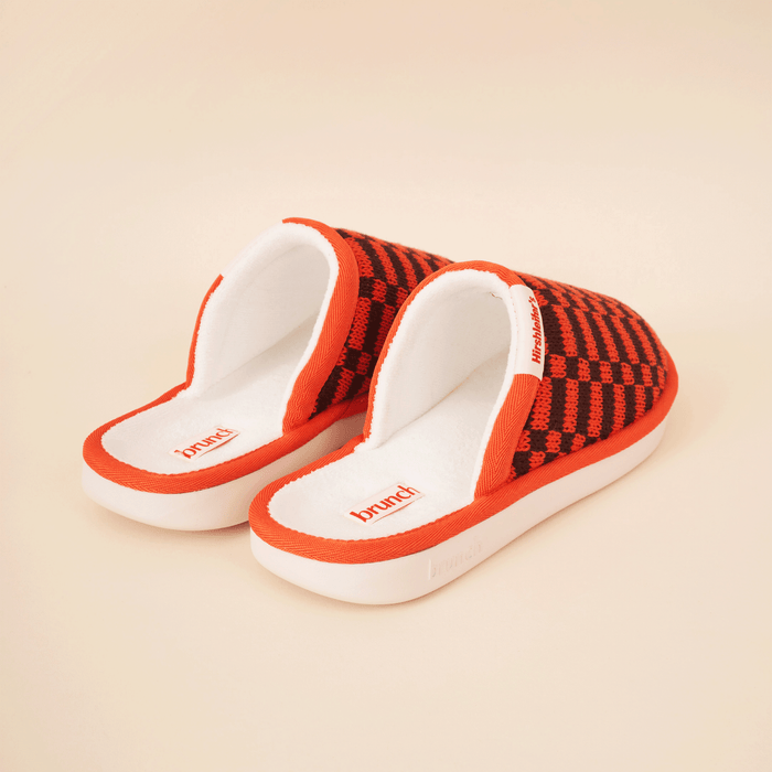 Three-quarter view of Amber Hirshleifers slipper with orange-black knit upper and white cushioned footbed on cream background