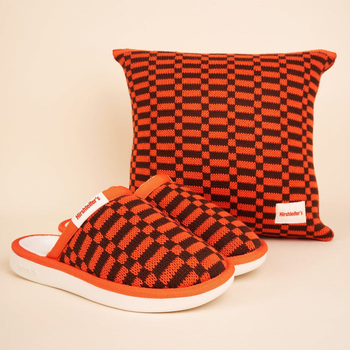 Three-quarter view of Amber Hirshleifers knit slippers showing orange and black checker knit upper and white foam sole on beige background