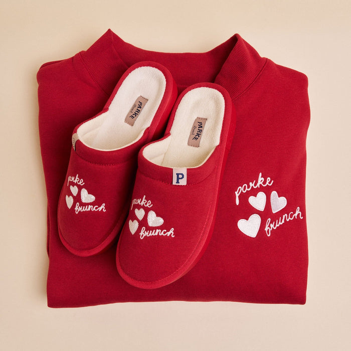 Top-down view of folded crimson Parke mockneck fleece with matching slippers embroidered 'parke' and 'brunch' with three hearts on a pale beige background