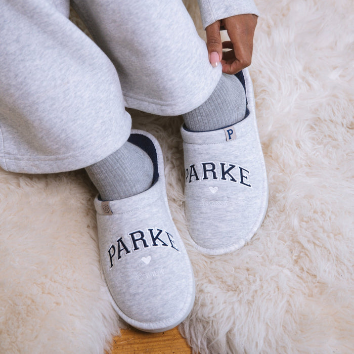 Top-down view of light grey Parke fleece slippers with embroidered PARKE lettering and slip-on design on a white rug