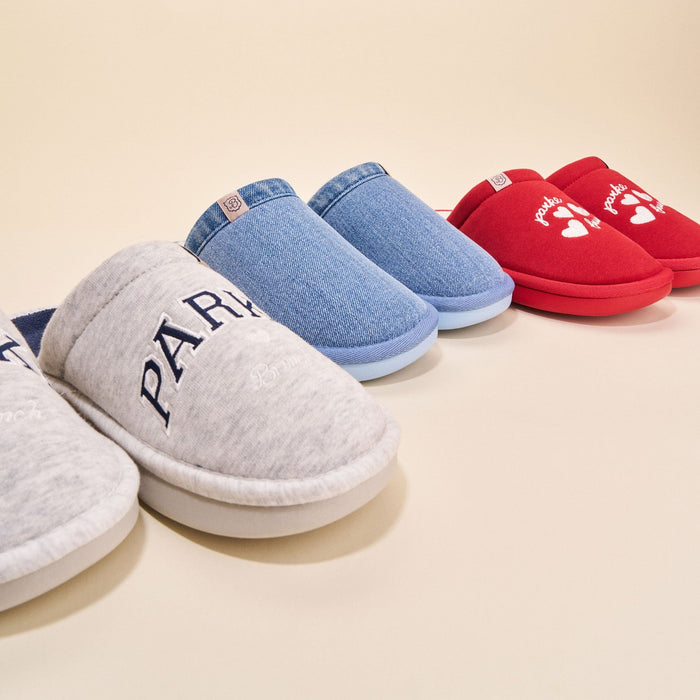 Three-quarter front view of Essential Parke Varsity fleece slippers: gray pair with PARKE embroidery, denim-look blue pair, and red pair with white-heart motif on beige background