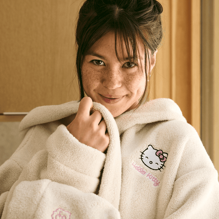 Front view of cream Essential Robe on model showing embroidered Hello Kitty chest, hood, oversized fit, and terry texture
