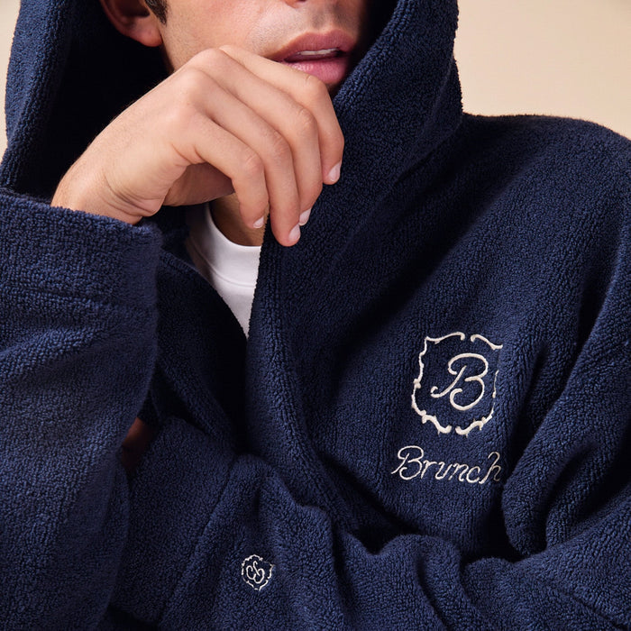 Close-up of navy Essential Robe on model showing plush terry fabric, hood, and embroidered Brunch logo on beige background