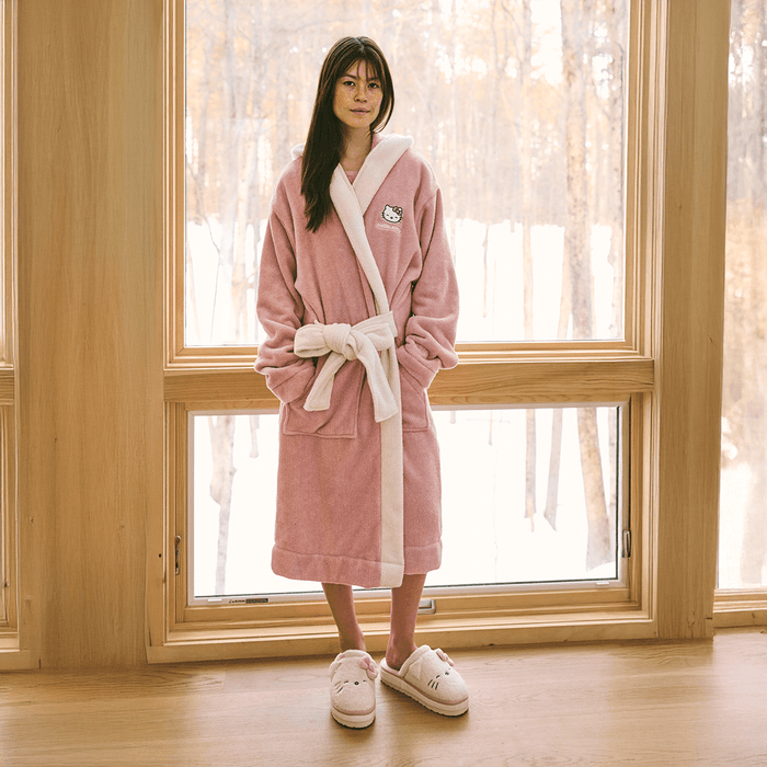 Front view of Essential Robe in pink showcasing embroidered Hello Kitty, cream trim, belt and pockets by a wood window