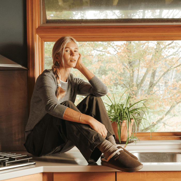 Woman seated on a kitchen counter wearing brown suede shearling slippers beside a potted plant and a window showing fall foliage