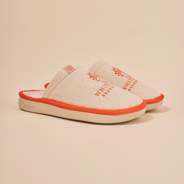 Angled view of The White Lotus slipper in beige with embroidered THE WHITE LOTUS RESORT, linen upper and orange trim