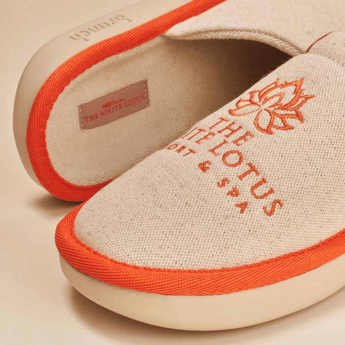 Close-up of The White Lotus slipper showing beige linen upper with orange embroidered lotus logo, orange piping and cushioned EVA sole
