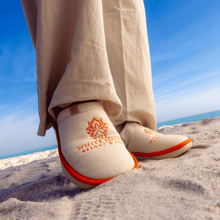 Low-angle front view of Essential The White Lotus slipper in beige showing orange embroidered lotus logo and EVA outsole on sand