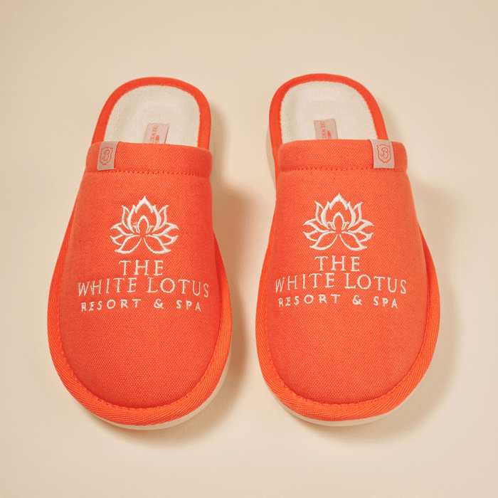 Top view of pair of orange White Lotus Essential slippers showing white embroidered 'THE WHITE LOTUS RESORT & SPA' logo and terry lining on a beige background