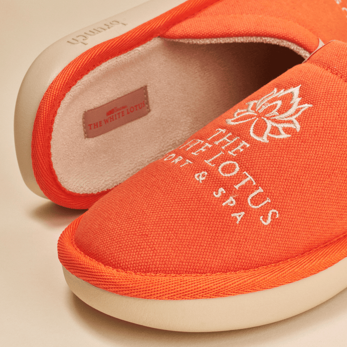 Close-up of orange The White Lotus slipper showing embroidered THE WHITE LOTUS Resort and Spa logo, linen upper and cushioned EVA sole on beige background
