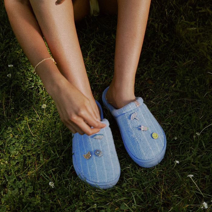 Angled top view of Cabana Essential XXL striped terry slippers showing blue-and-white stripes and attached charms on grass