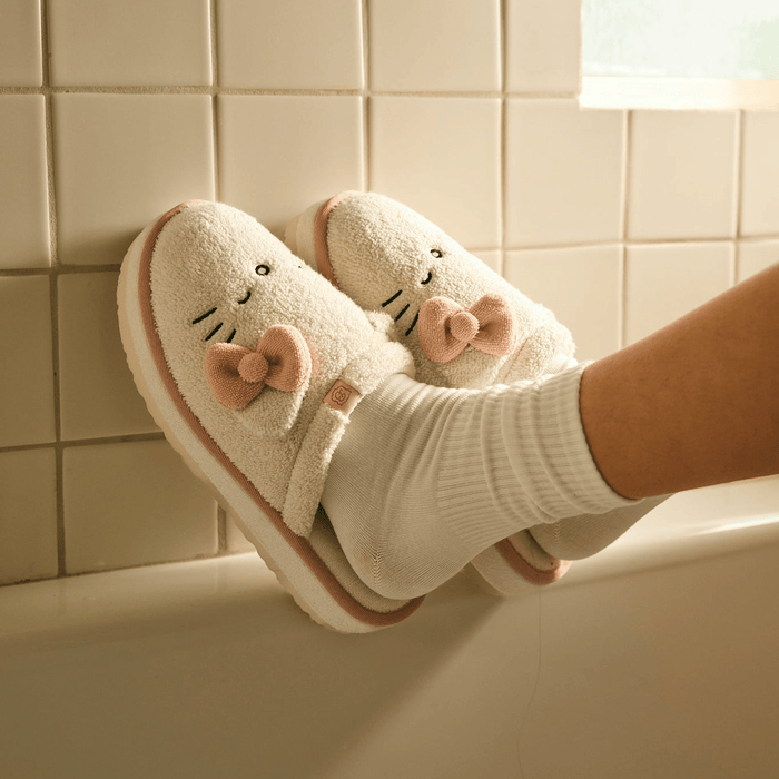 Angled side view of Essential XXL Hello Kitty cream terry slippers on feet showcasing 3D ears and pink bow with tiled bathtub background