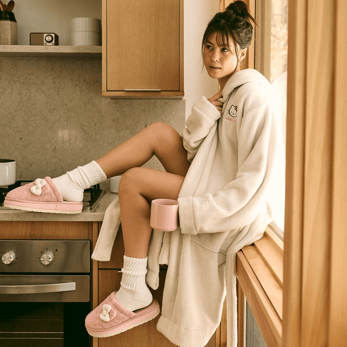 Woman in a cream robe sitting on a kitchen counter wearing pink Hello Kitty terry slippers with white bow and holding a pink mug