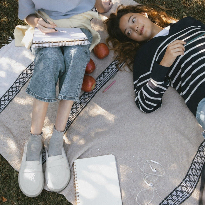 Person wearing cream suede Yankees loafers sitting with legs extended on a picnic blanket with notebook, apples, and earbuds nearby