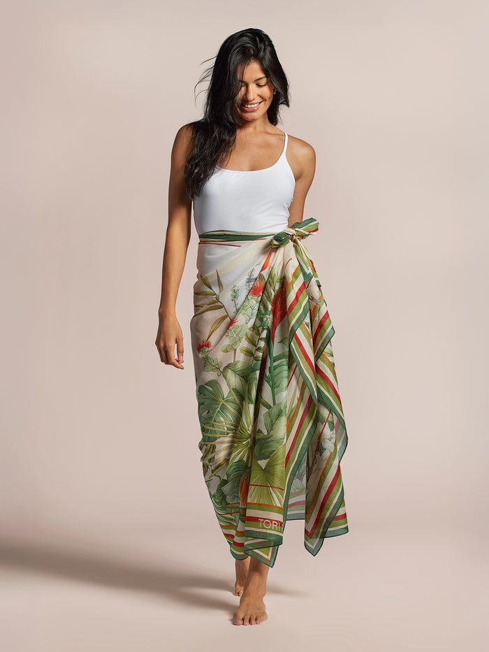 Woman in white tank top and green floral-print Manu Garden pareo, barefoot, looking down, front view.