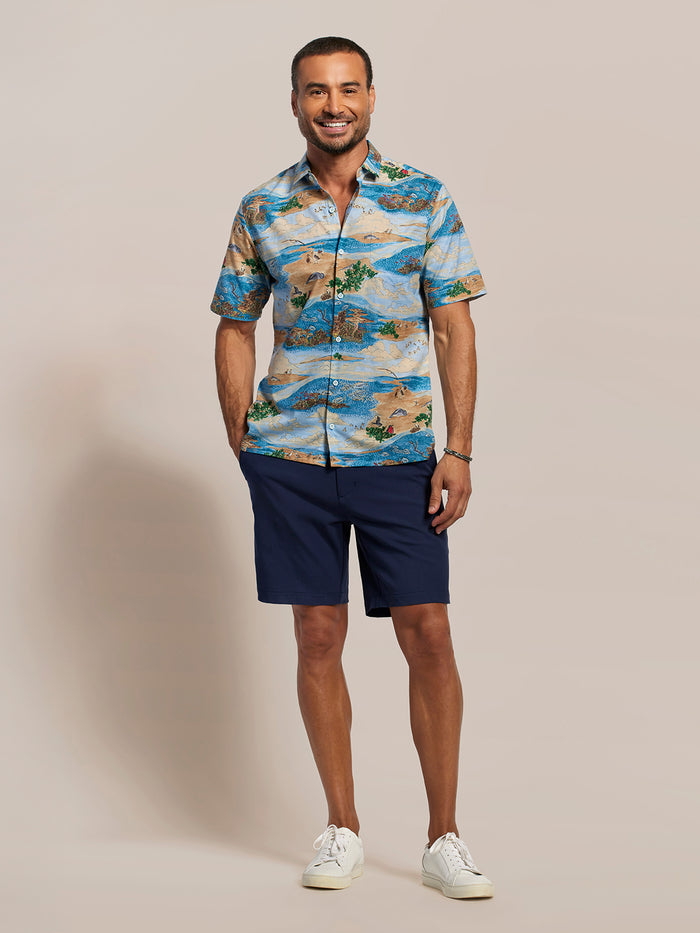 Ocean Blue, green, and beige island-print cotton short-sleeve Hawaiian shirt and navy shorts, front view of a man, plain background.