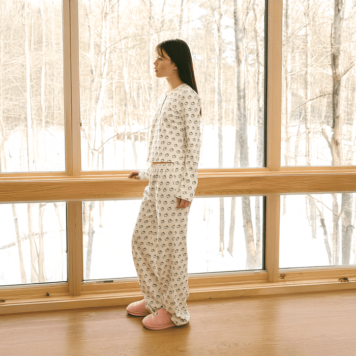 Side view of cream Hello Kitty cotton pajama set on model showcasing all-over Hello Kitty print and snap-front long-sleeve top by a snowy window