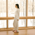 Side view of cream Hello Kitty cotton pajama set on model showcasing all-over Hello Kitty print and snap-front long-sleeve top by a snowy window